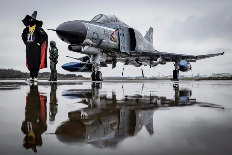 A person in a black and red costume with a yellow face mask and a tall black hat stands next to a gray military jet on a wet…
