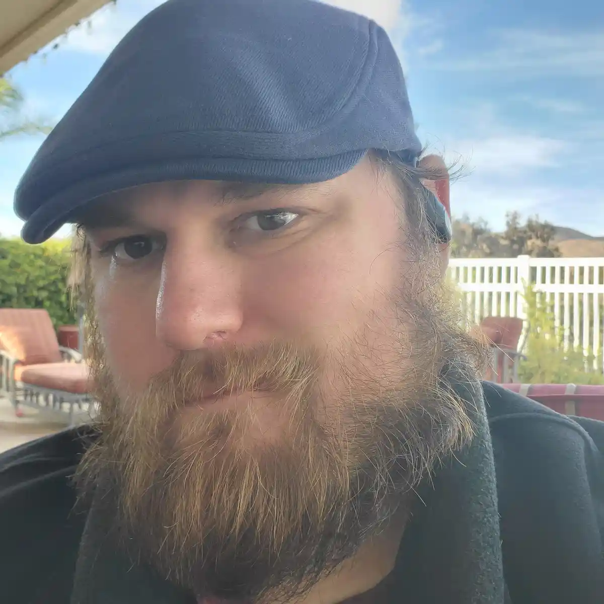 A close-up selfie of a man with a full beard and a blue flat cap, looking directly at the camera with a serious expression…