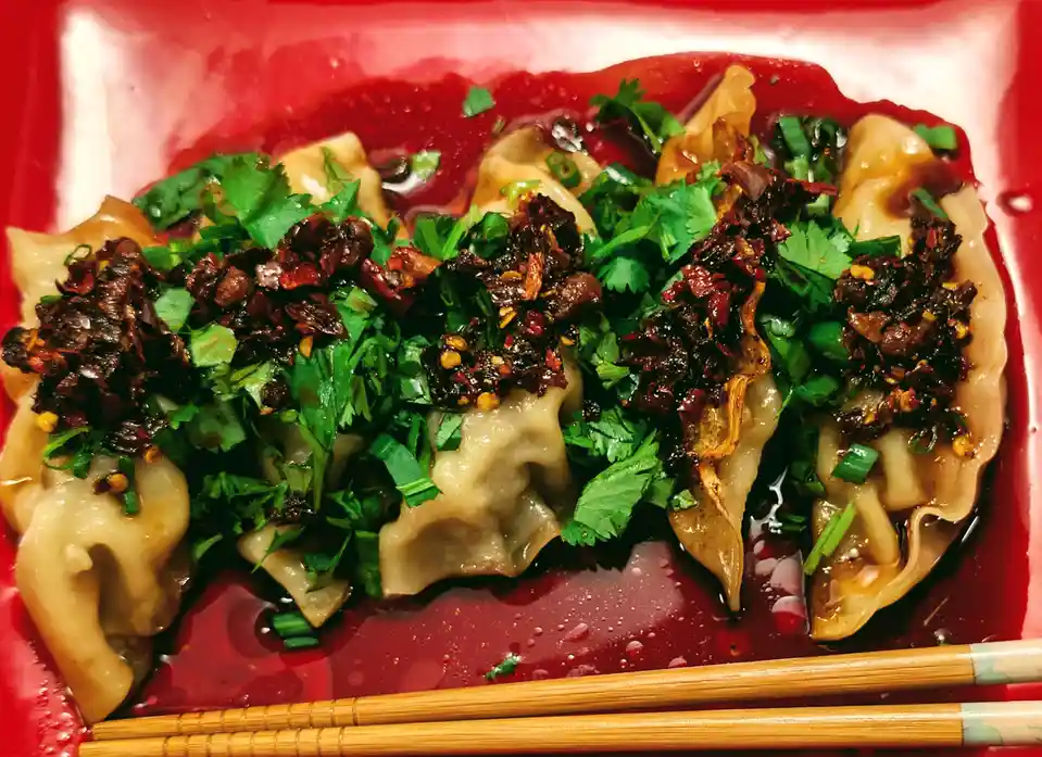 A close-up photo shows several dumplings in a red tray, covered in a dark sauce, chopped green herbs, and fried chili oil. A…
