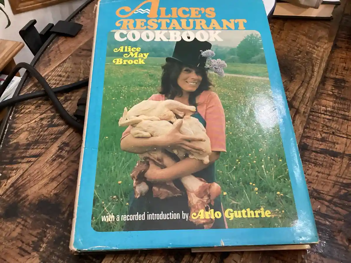 A physical copy of 'Alice's Restaurant Cookbook' by Alice May Brock, featuring a photo of a woman in a top hat holding raw…
