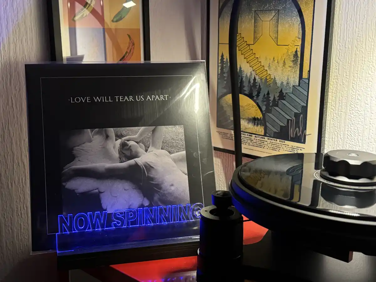 A framed vinyl record cover for Joy Division's 'Love Will Tear Us Apart' sits next to a turntable, with a glowing blue neon…