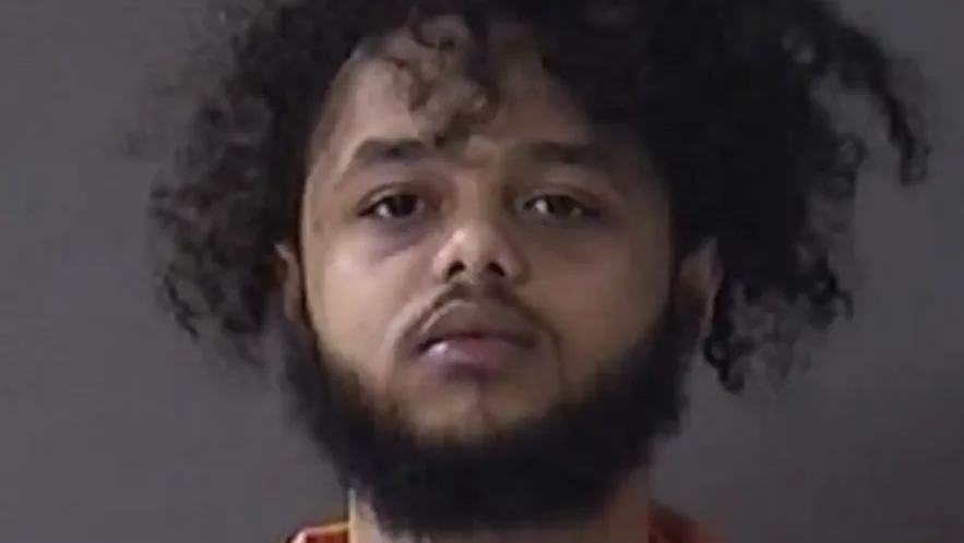 A mugshot of a man with dark curly hair and a beard, wearing an orange shirt, stares blankly at the camera. Overlaid text…