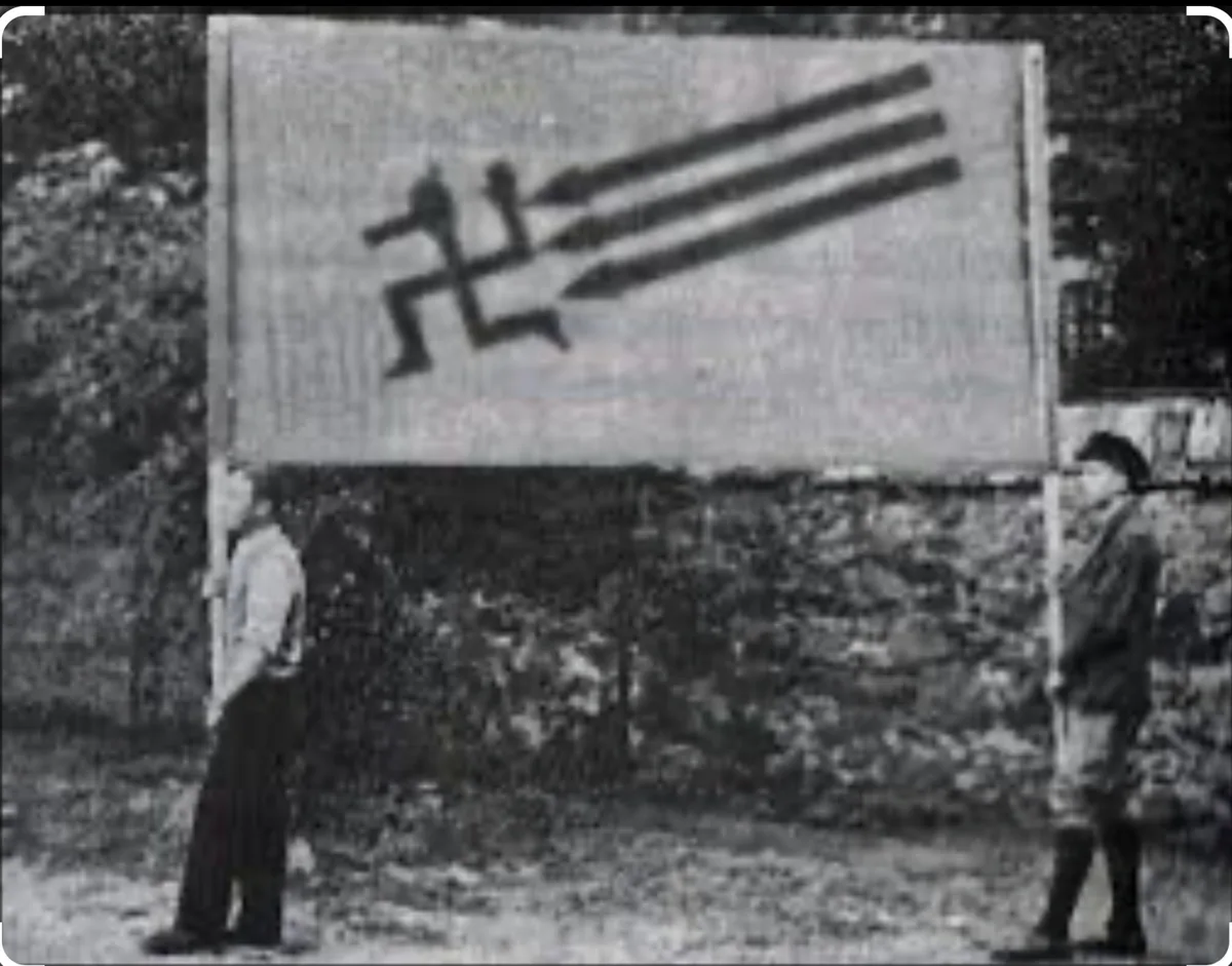 A grainy black-and-white photo shows two men standing on either side of a large banner displaying a swastika with three…