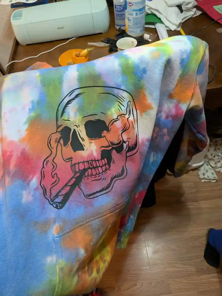 A tie-dye hoodie with a black outline of a skull holding a cigar is draped over a chair. The background shows a cluttered…