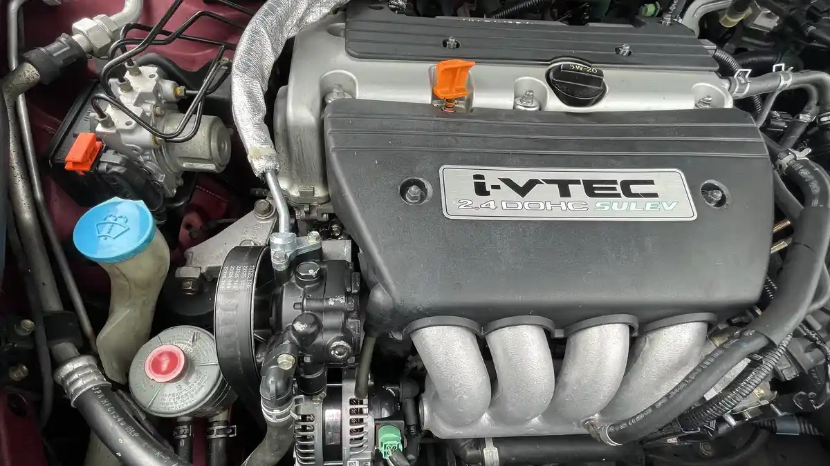 A close-up photo of a car engine bay showing a black engine cover with a silver plaque that reads 'i-VTEC 2.4 DOHC SULEV'…