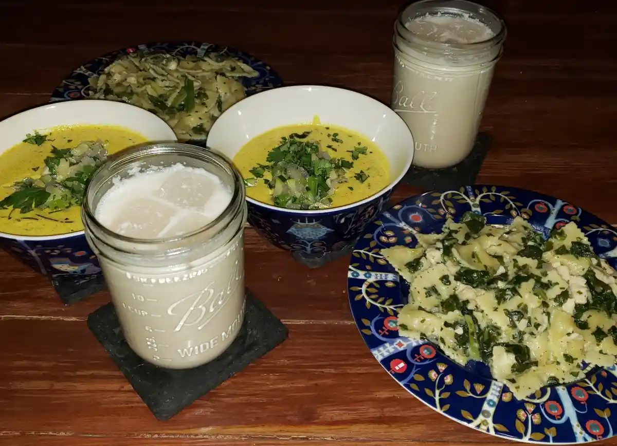 A wooden table displays two bowls of yellow curry soup topped with chopped green herbs, two plates of pasta with greens, and…