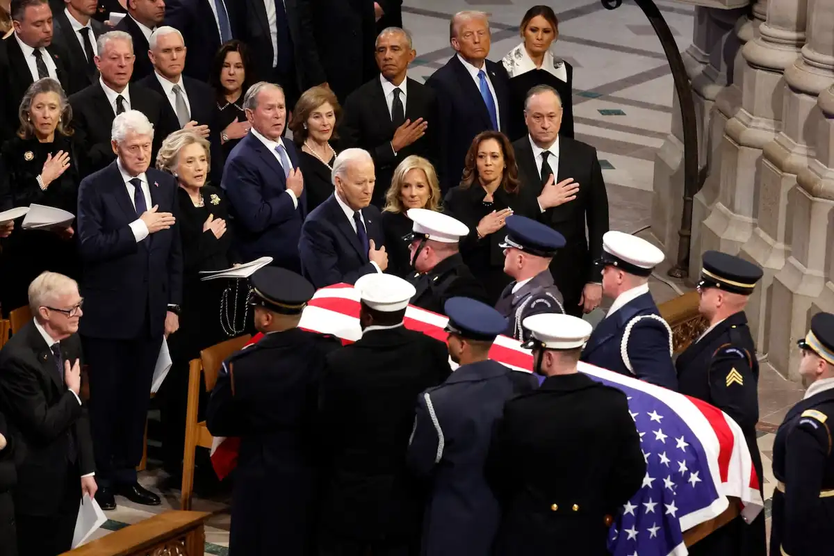a solemn photo of former U.S. presidents and their families standing in a church during a funeral, with military personnel…