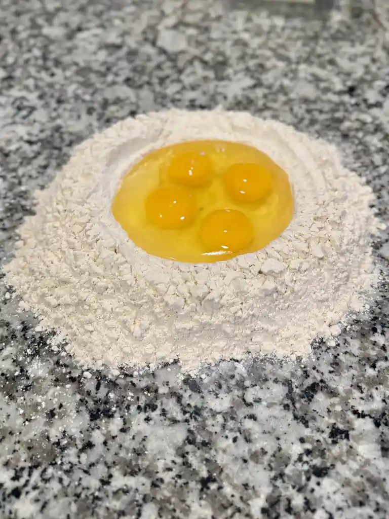 A close-up photo shows four cracked egg yolks sitting in a circular well of white flour on a speckled granite countertop…