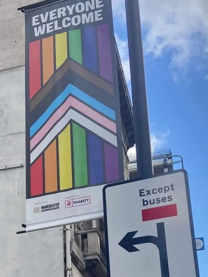 A large rainbow-patterned banner on a building reads 'EVERYONE WELCOME' with logos for Manchester Accommodation and a…