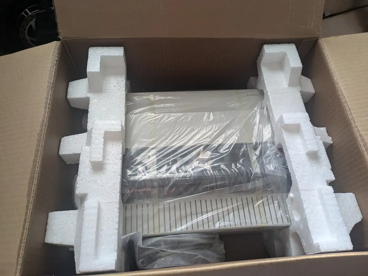 An overhead shot of a cardboard box containing a printer wrapped in plastic, surrounded by white styrofoam inserts. The text…
