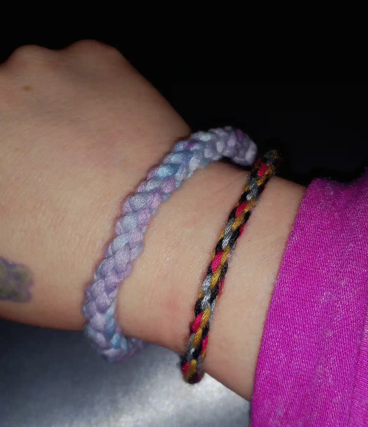 A close-up of a person's wrist wearing two braided friendship bracelets: one is a pastel purple and blue, the other is…