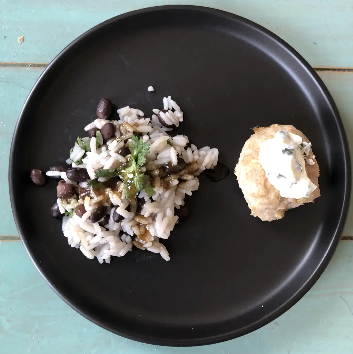 A black plate holds a small serving of rice mixed with black beans and cilantro, alongside a single round piece of bread…