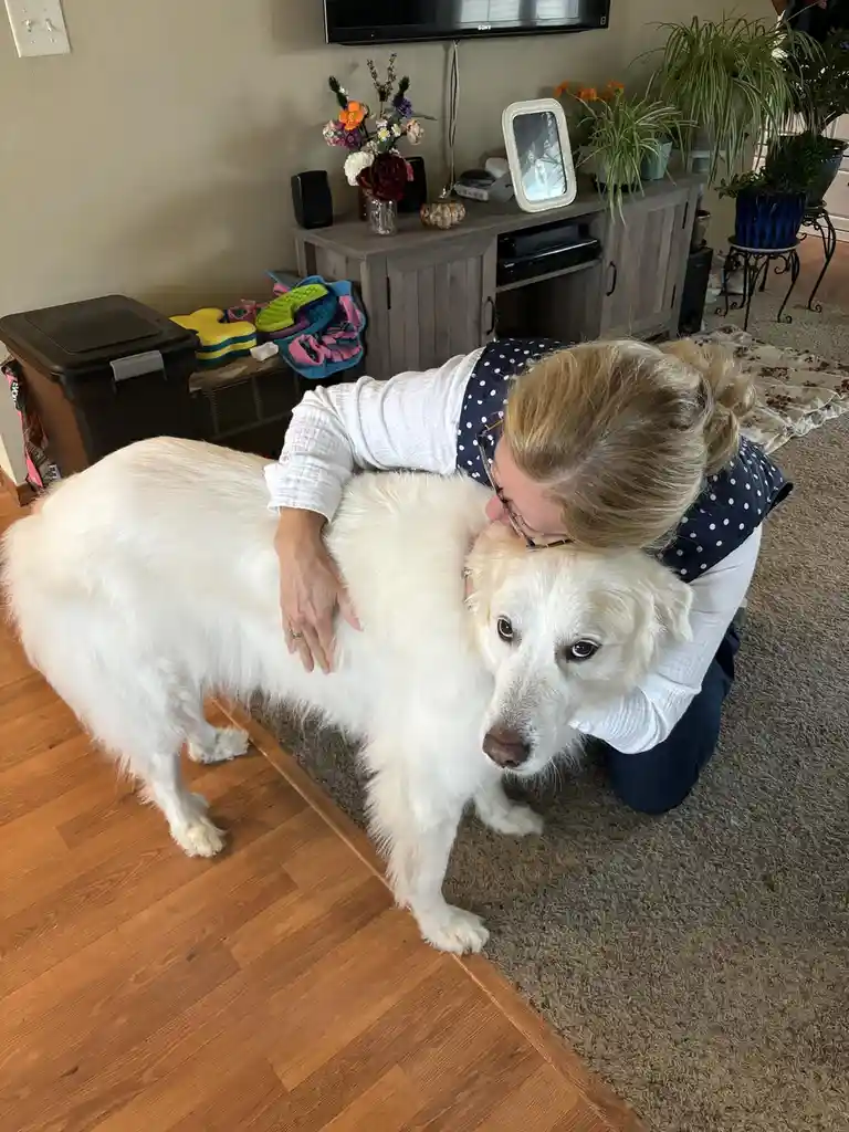A woman with blonde hair and glasses is kneeling on a carpeted floor, hugging a large, fluffy white dog from behind. The…