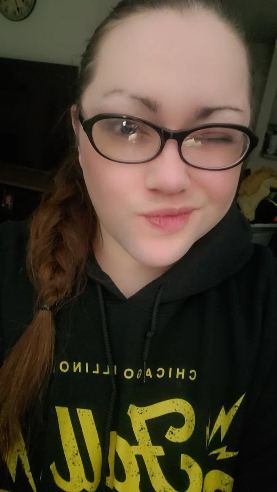 A person with glasses and a braid is winking and smiling while wearing a black hoodie with yellow text that reads 'CHICAGO…