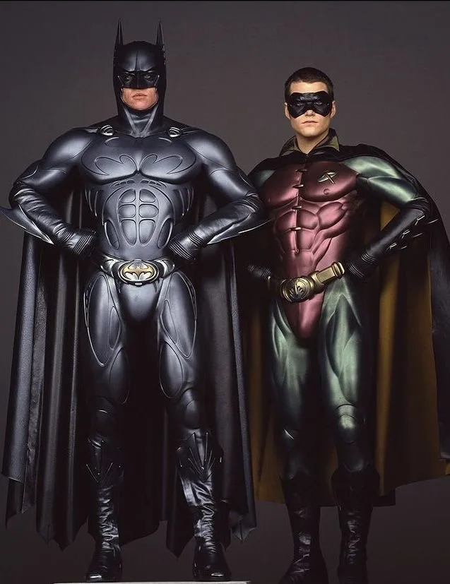 Two men in full superhero costumes stand side by side against a plain gray background. The man on the left wears a black…