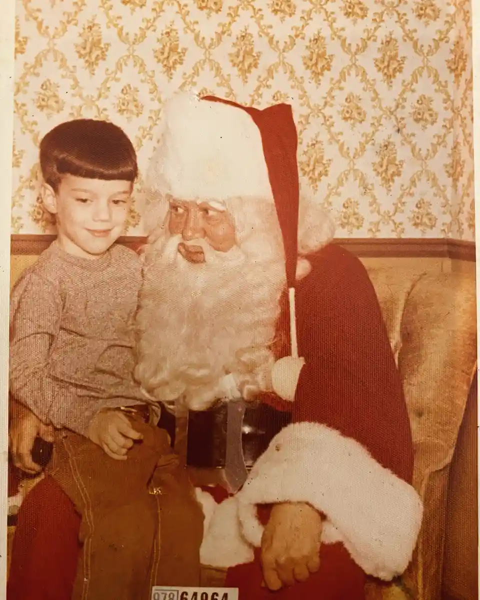 A vintage, sepia-toned photo shows a young boy sitting beside a large, traditional Santa Claus figure on a couch. The boy…