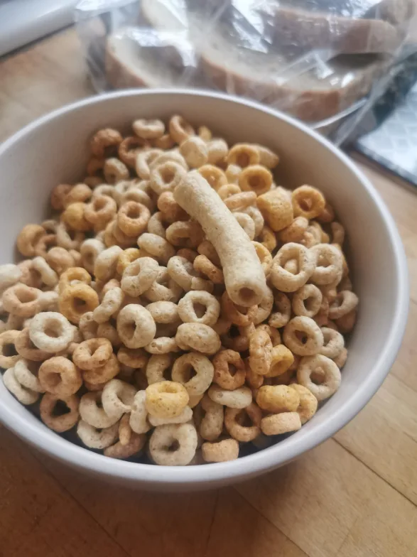 A close-up photo shows a white bowl filled with cereal rings and one long, cylindrical cereal piece standing upright among…
