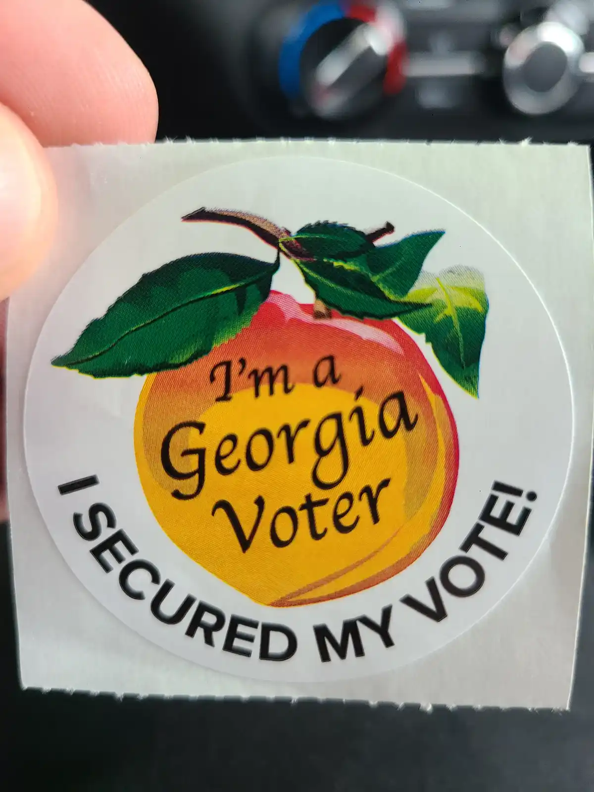 A close-up photo shows a hand holding a circular sticker featuring a peach with green leaves. The sticker has text inside…
