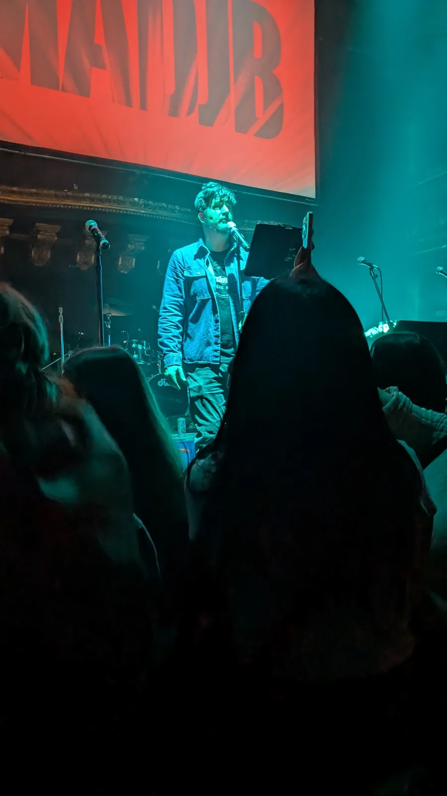 A man with a beard and denim jacket stands on a dimly lit stage under green lighting, singing into a microphone while…