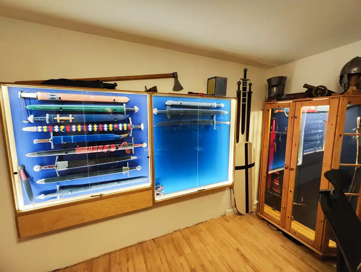 A room with wooden floors and beige walls displays an extensive collection of swords and medieval-style weapons. Two large…