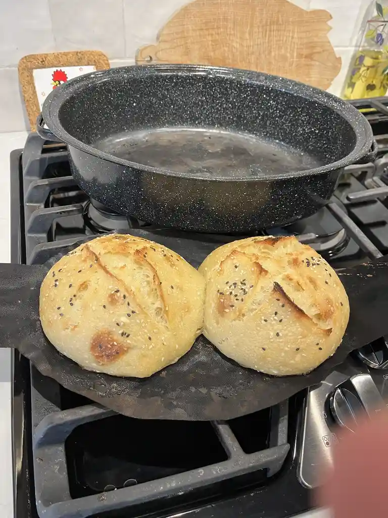 Two round, golden-brown bread rolls with sesame seeds sit side-by-side on a black baking sheet on a stovetop, appearing to…