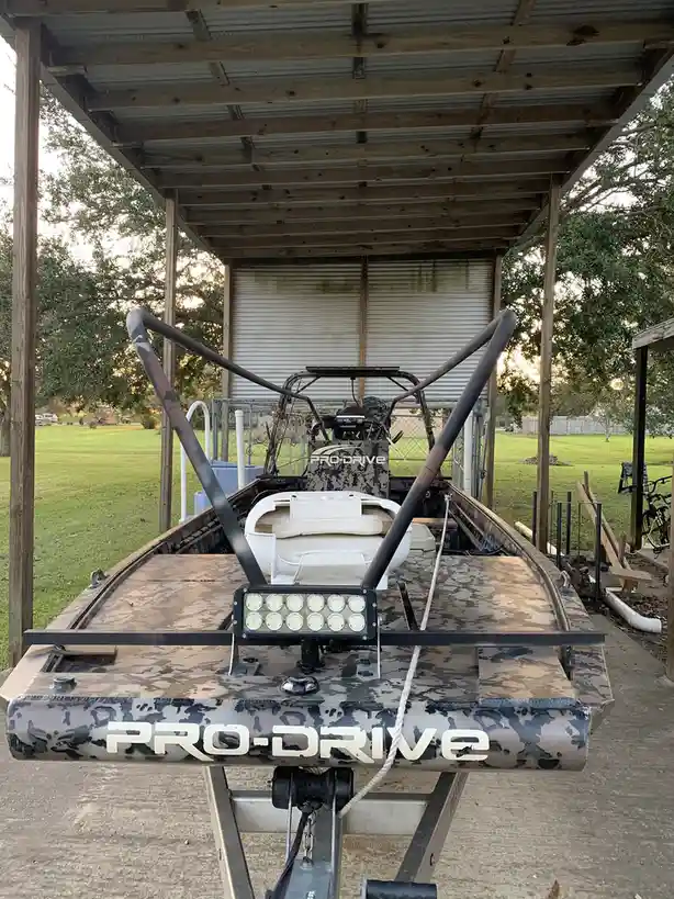 A camouflage-patterned Pro-Drive boat is parked under a wooden shelter on a concrete pad, with a white seat and a light bar…