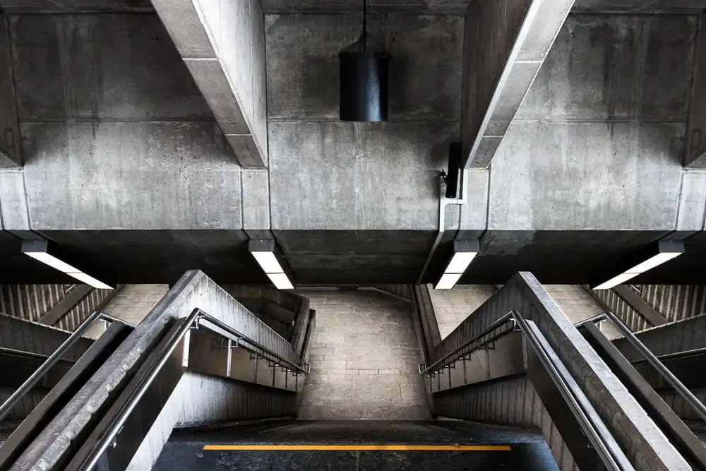 A low-angle photo looking up at a concrete subway or underground station staircase with metal railings and fluorescent…