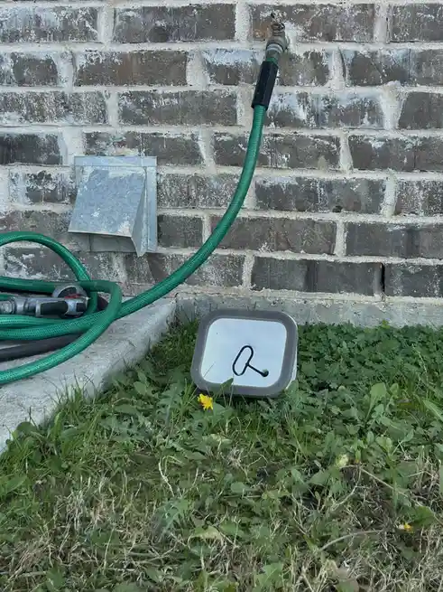A green garden hose is connected to an outdoor spigot on a brick wall, with its nozzle and coiled hose resting on a concrete…