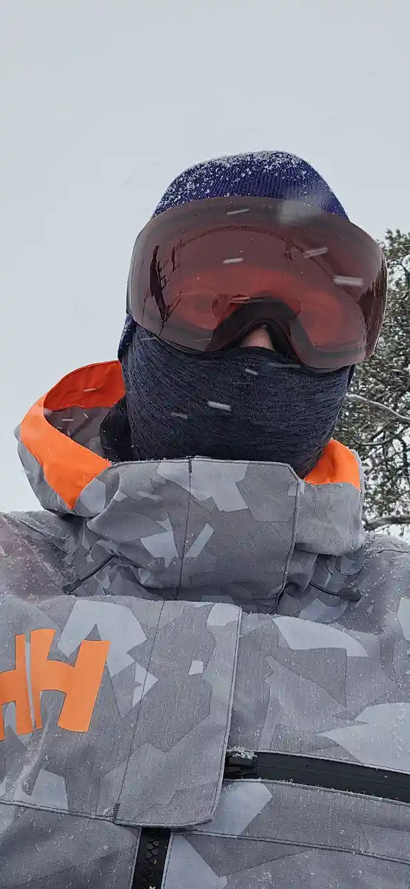 A person in a snowy environment wears a blue beanie, a black balaclava, and large orange-tinted ski goggles, with only their…
