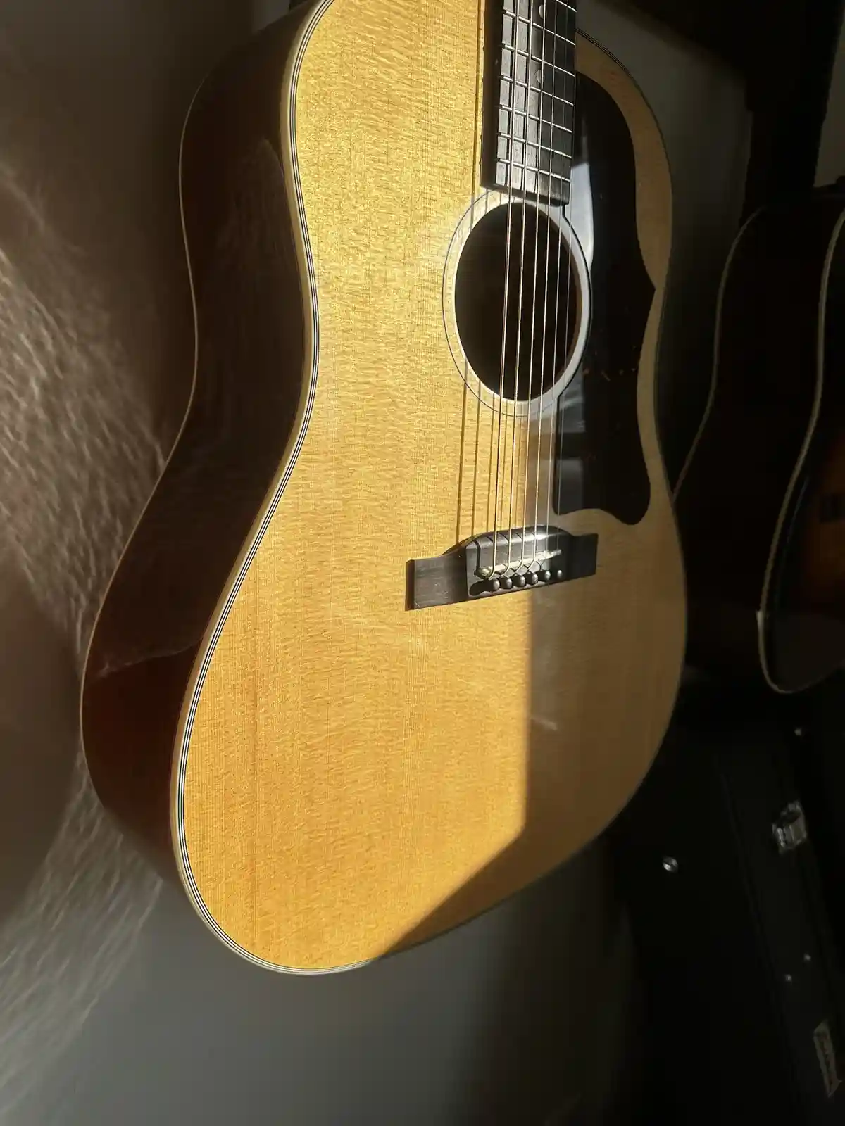 A close-up photo of a light-wood acoustic guitar with a dark brown back and binding, bathed in warm sunlight that highlights…