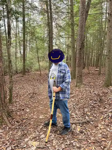 A person stands on a leaf-covered forest trail, wearing a blue plaid shirt over a gray t-shirt with the text 'their fight is…