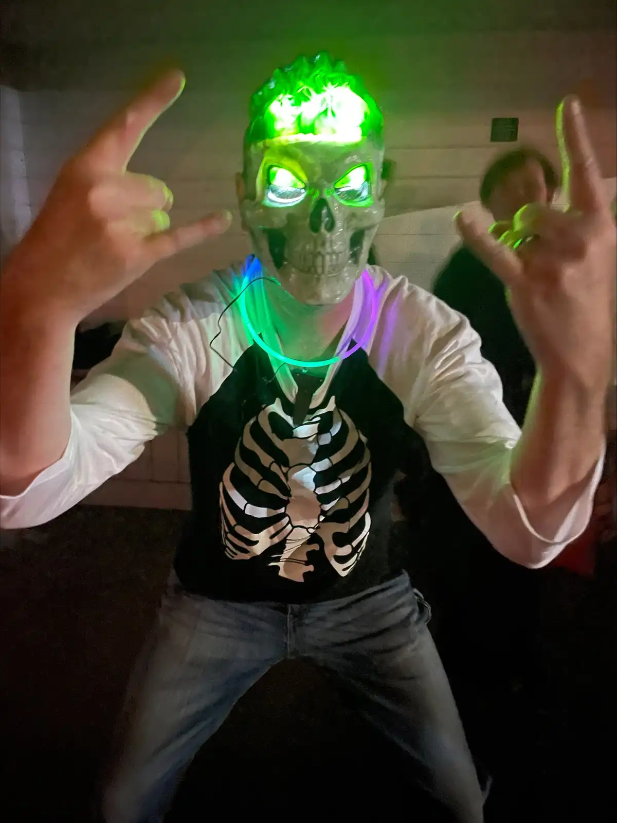 A person wearing a glowing green skull mask and a black vest with a skeleton chest design poses with both hands making…
