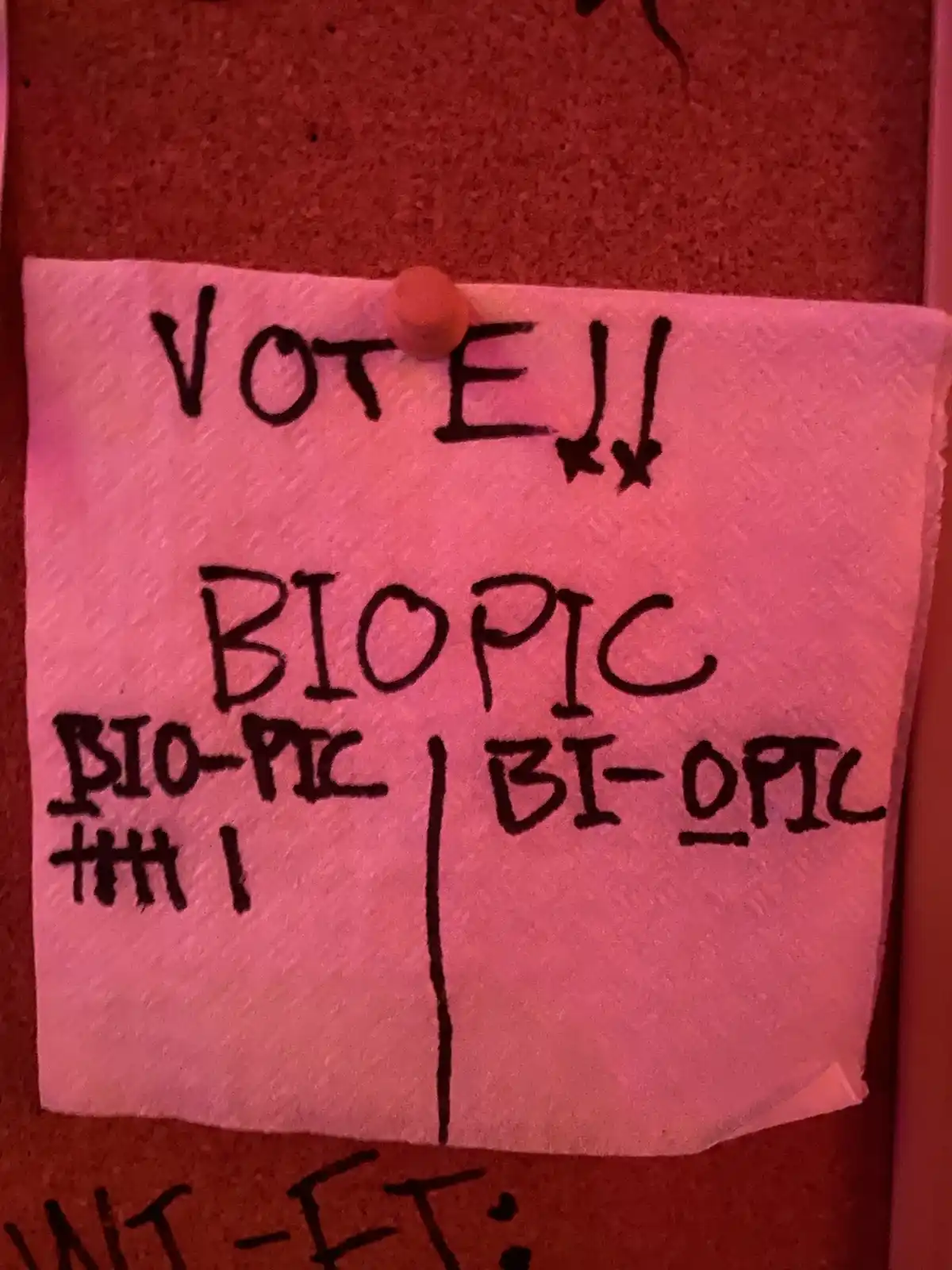 A handwritten note on a paper towel pinned to a corkboard urges viewers to vote between two options: 'BIOPIC' (with arrows…