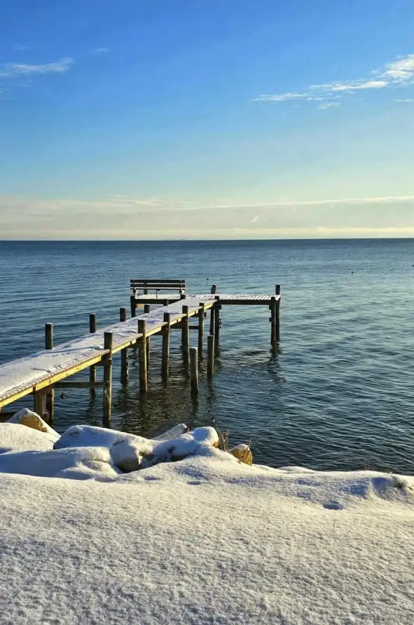 A serene winter scene shows a wooden pier extending into calm water, covered in snow, with a snow-dusted shoreline in the…