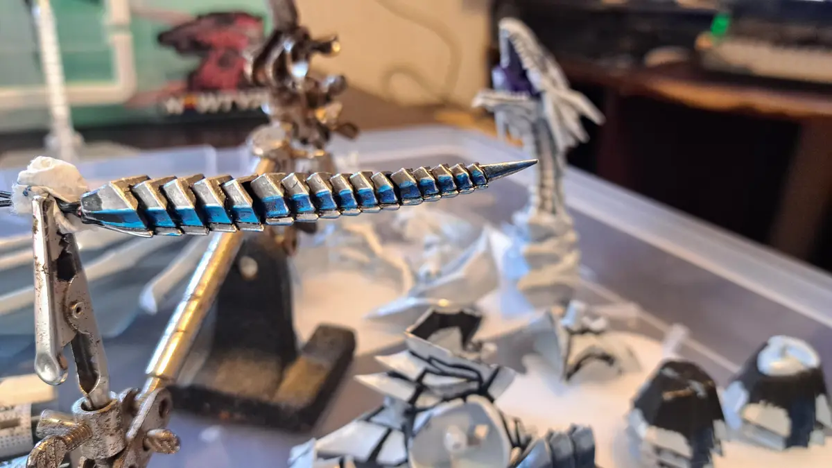 A close-up photo shows a metallic, segmented, spiked weapon with blue painted edges, held in a clamp on a workbench. In the…
