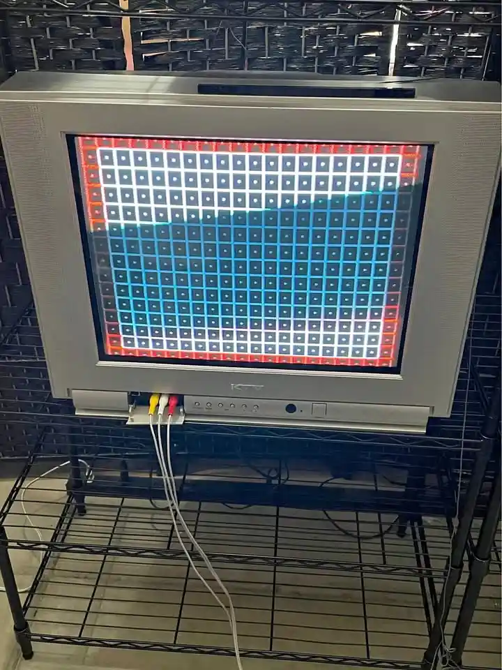 An old silver CRT television sits on a black metal shelf, its screen displaying a grid of red, white, and blue squares…