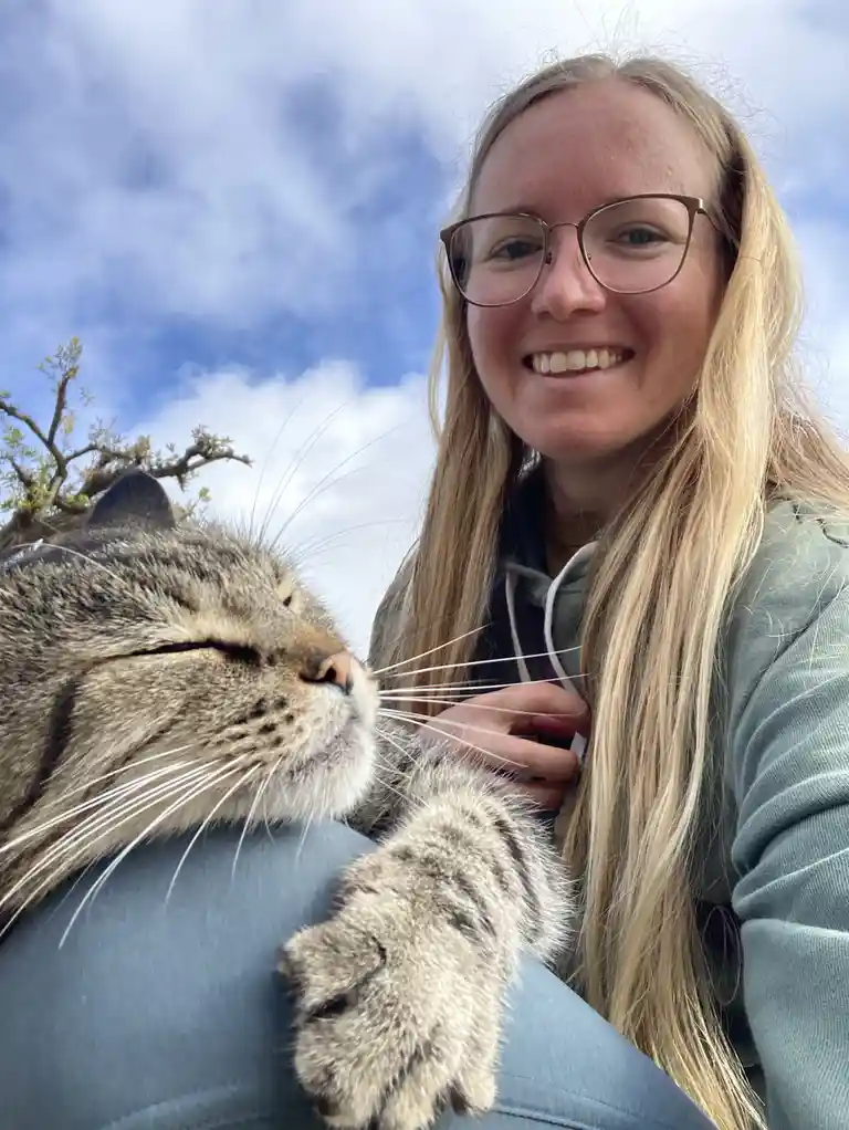 A smiling woman with blonde hair and glasses holds a content tabby cat that has its eyes closed and paw resting on her lap…