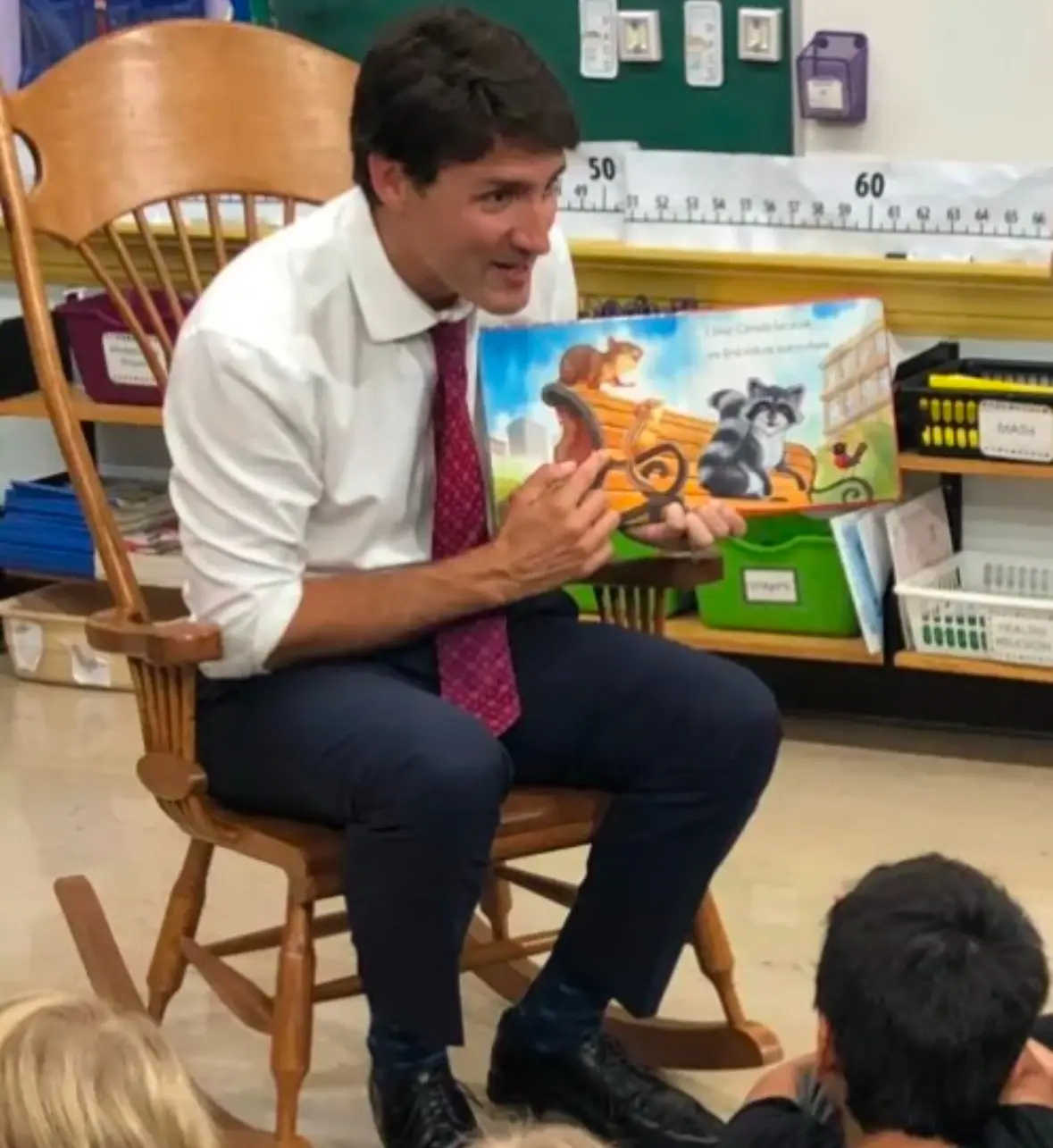 A man in a white shirt and red tie sits in a wooden rocking chair in a classroom, holding up a children's book with cartoon…