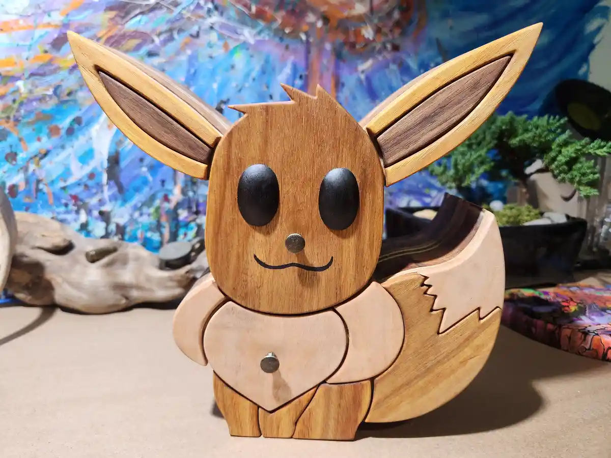 A wooden sculpture of the Pokémon Eevee, crafted from various wood tones with black eyes and a smiling face, sits on a…