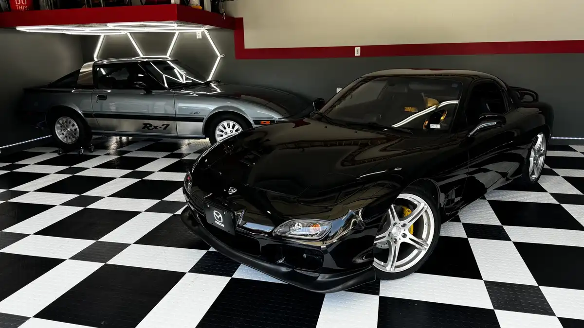 Two Mazda RX-7 sports cars are displayed in a garage with a black-and-white checkered floor. The foreground features a…