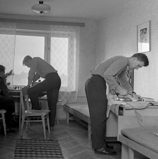 A black-and-white photo shows three men in a sparsely furnished room. One man stands on a chair, leaning over a table, while…