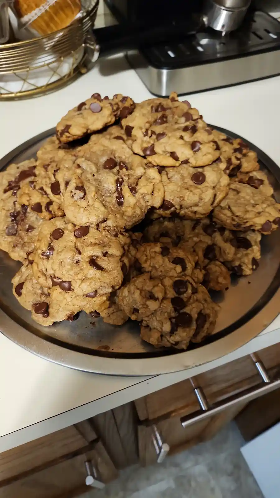 A large metal tray piled high with freshly baked chocolate chip cookies sits on a kitchen counter, with a coffee machine and…