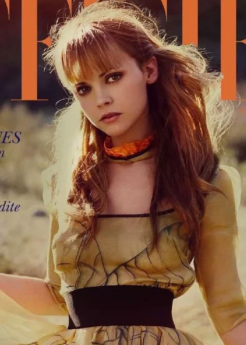 A woman with long brown hair and bangs stares directly at the camera with a neutral expression, wearing a yellow dress with…