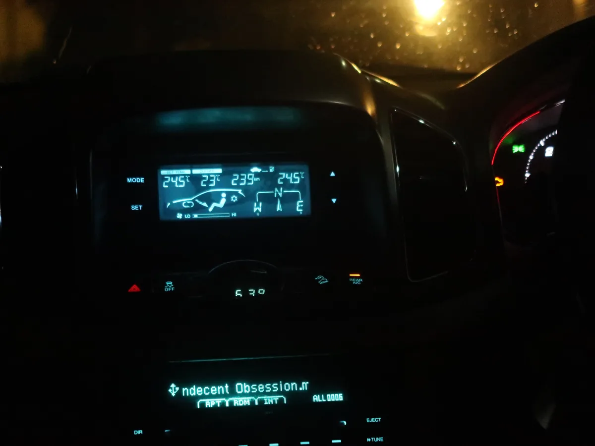 A nighttime car interior shot showing a digital dashboard with the temperature set to 24.5°C, outside temperature 23°C, and…