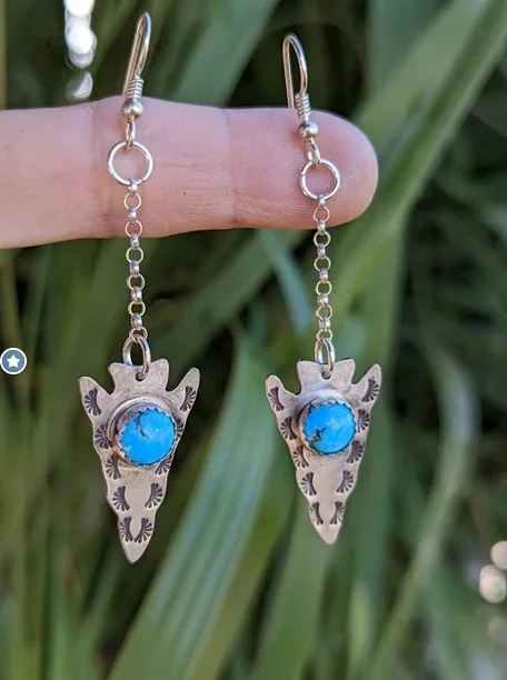 A hand holds a pair of silver dangle earrings shaped like arrowheads, each featuring a bright blue turquoise stone in the…