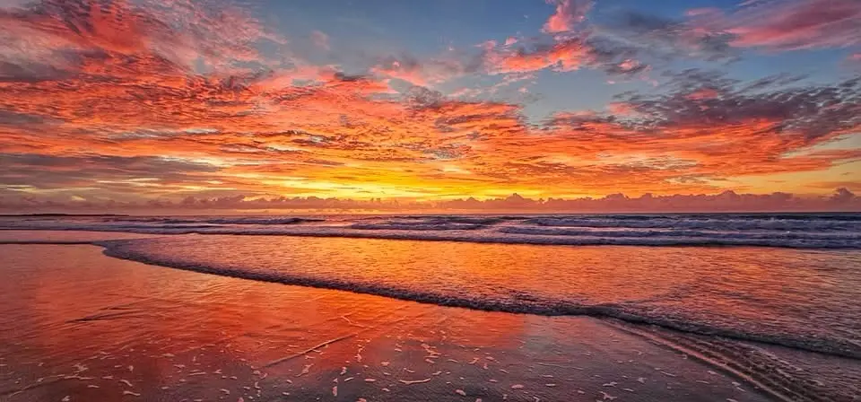 A panoramic photo of a vibrant sunset over a beach, with fiery orange and pink clouds reflecting on wet sand and gentle…