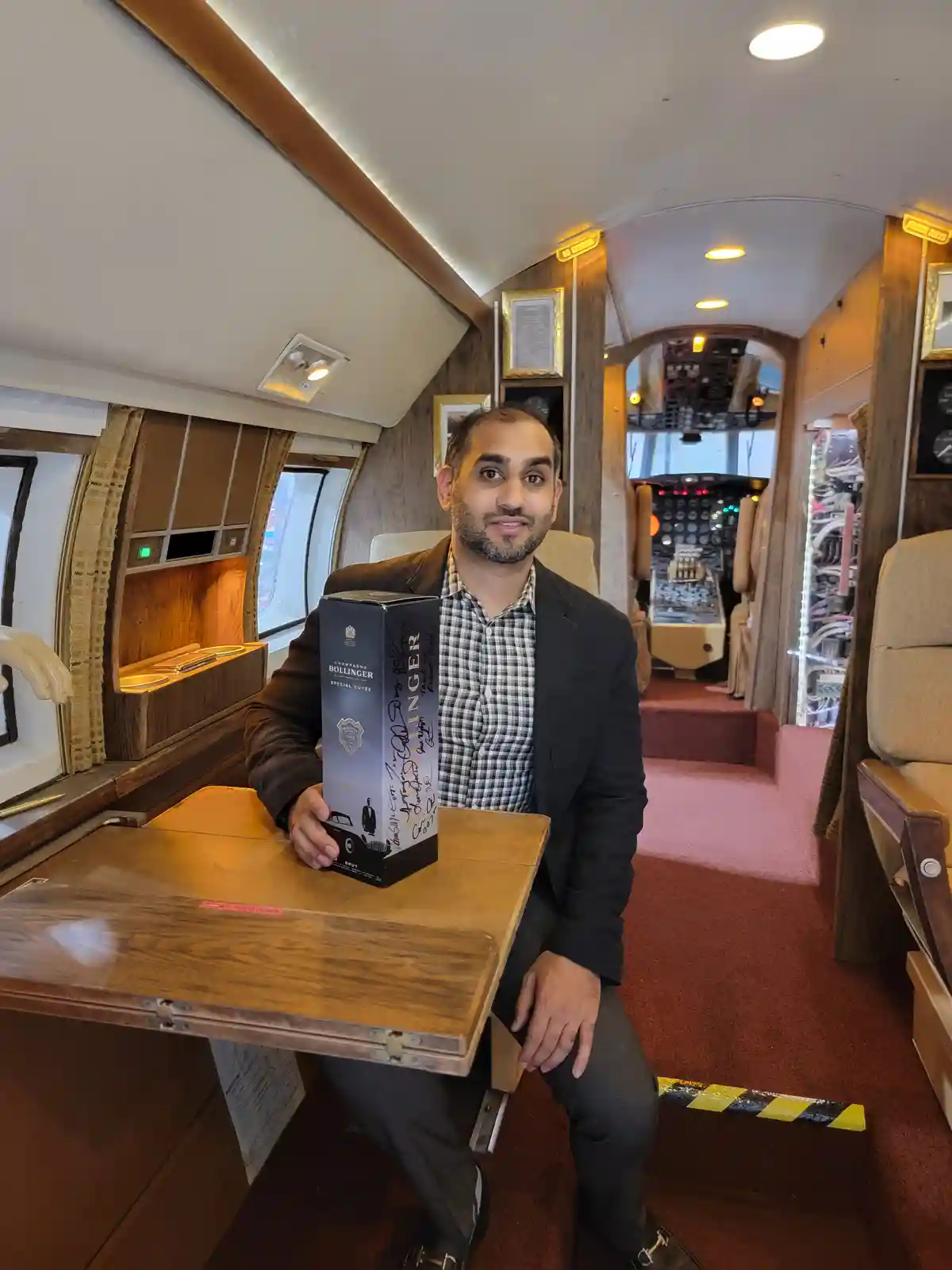 A man in a suit sits inside a luxurious private airplane cabin, holding a tall box of Bollinger champagne. The cabin…