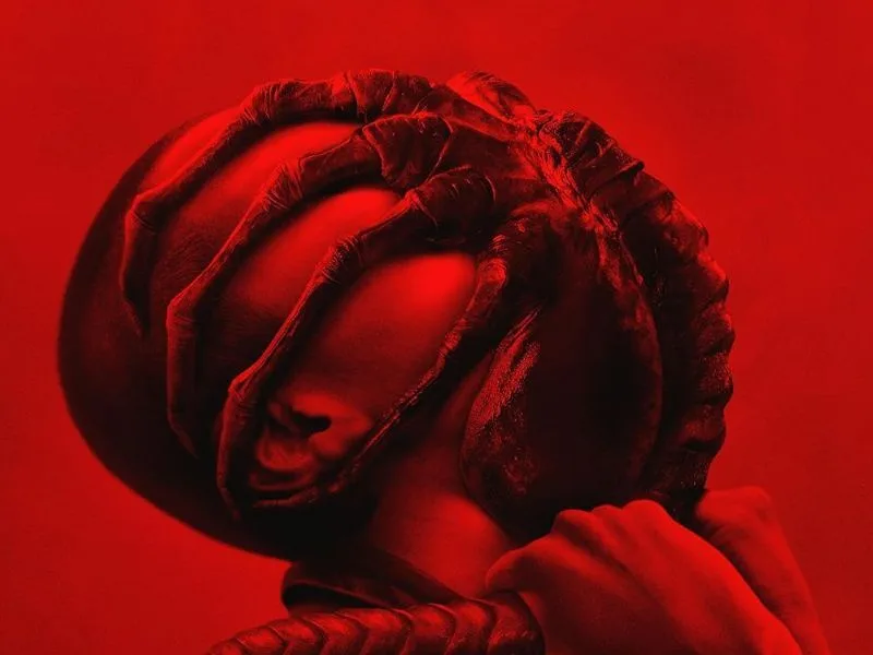 A person's face is obscured by a large, textured, red-colored object resembling a grotesque, alien-like hand or tentacle…