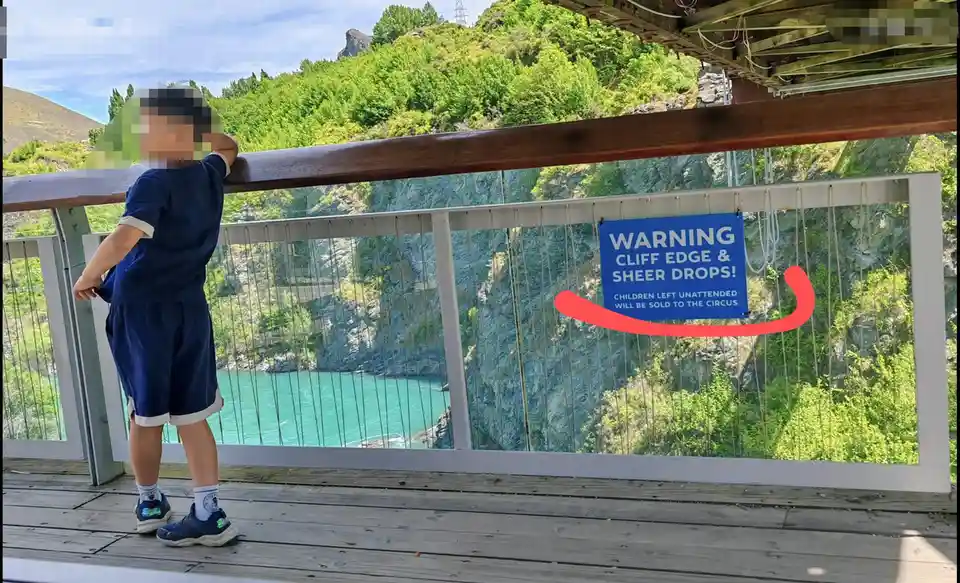 A young child in a blue uniform leans against a railing on a wooden bridge overlooking a steep, green canyon with turquoise…
