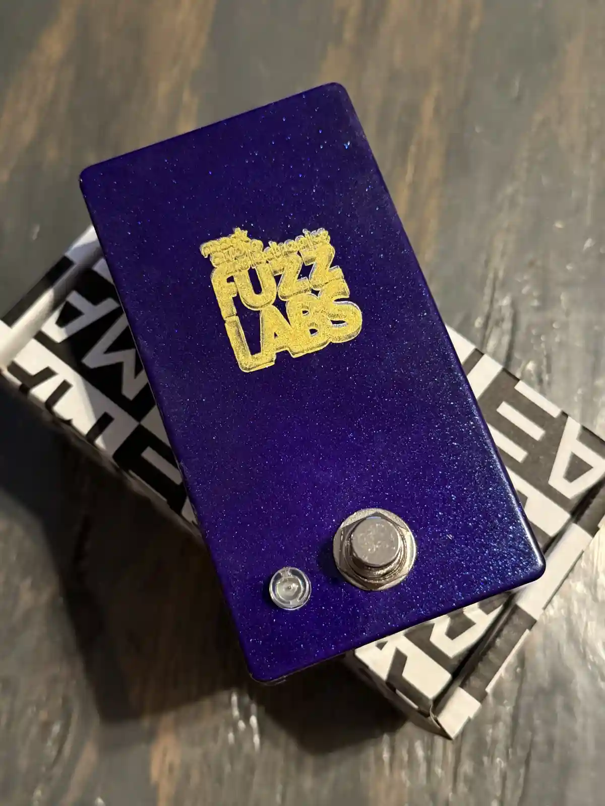 A close-up photo of a purple glitter-finished guitar effects pedal with the brand name 'The Fuzz Labs' in gold lettering…